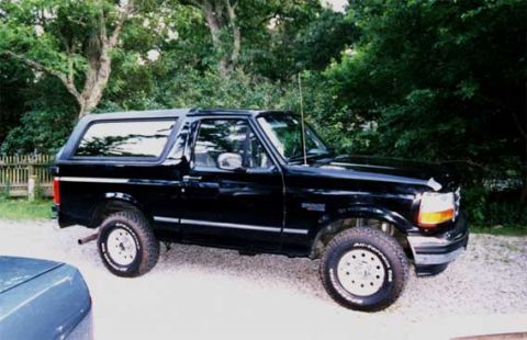 1996 Ford Bronco - This black 1996 Ford Bronco is seen from a three-quarter front angle, parked on a gravel surface with trees in the background.