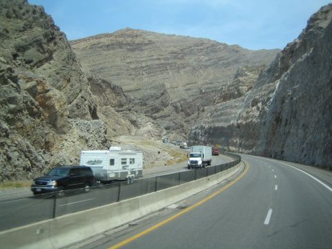 1990 Ford Bronco - black SUV tows a travel trailer along a canyon highway as a box truck follows beneath steep rock walls.
