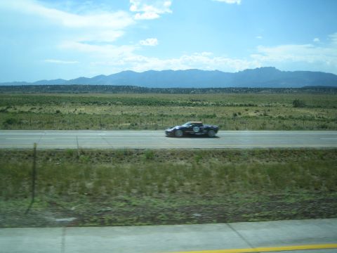 1990 Ford Bronco - dark sports car blurs along a desert highway across a grassy field with distant mountains.