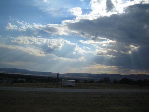 1990 Ford Bronco - Sunbeams pierce heavy clouds over a wide valley with a small white trailer parked in a field.