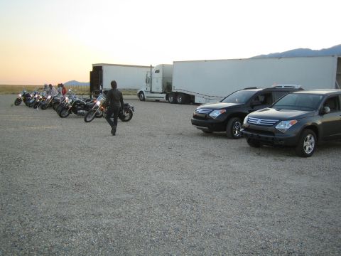 1990 Ford Bronco - row of cruiser motorcycles and dark SUVs sit on a gravel lot beside semi trailers at dusk.