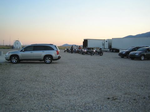 1990 Ford Bronco - silver crossover SUV and a cluster of motorcycles sit on a gravel lot near box trailers at dusk.