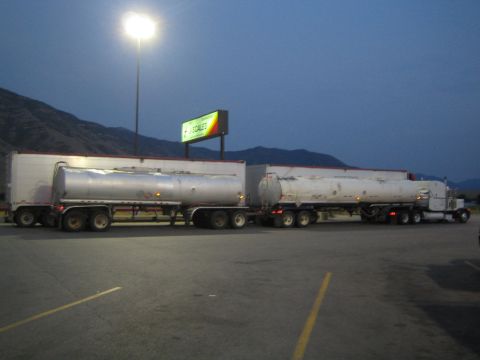 1990 Ford Bronco - semi truck with two silver tanker trailers sits in a truck-stop lot beneath a lit sign and mountains at dusk.