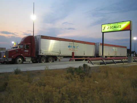 1990 Ford Bronco - red semi truck pulling two box trailers sits near a lit 'J-Scales' weigh-station sign at dusk.