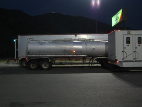 1990 Ford Bronco - silver tanker trailer is parked side-on in a lot at night, lit by overhead lights with hills behind.