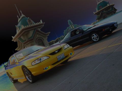 1998 Ford Mustang - Two Ford Mustangs are parked on a road with distinctive buildings in the background.