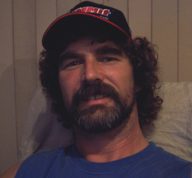 Man with beard and curly hair wearing a black baseball cap with red trim and a blue t-shirt