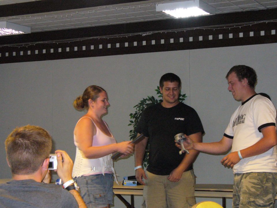 Woman in pink tank top and denim shorts accepts an award from a man in a black t-shirt; another man in a white t-shirt looks on