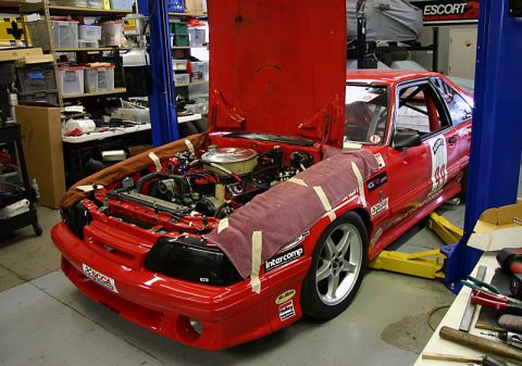 1993 Ford Mustang - A red 1993 Ford Mustang race car is undergoing maintenance in a workshop, with its hood open and engine visible.