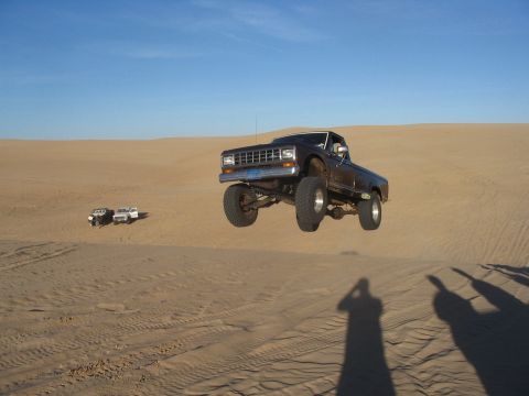 1985 Ford Ranger - A lifted pickup truck soars through the air against a clear blue sky, with other vehicles visible in the sandy landscape.