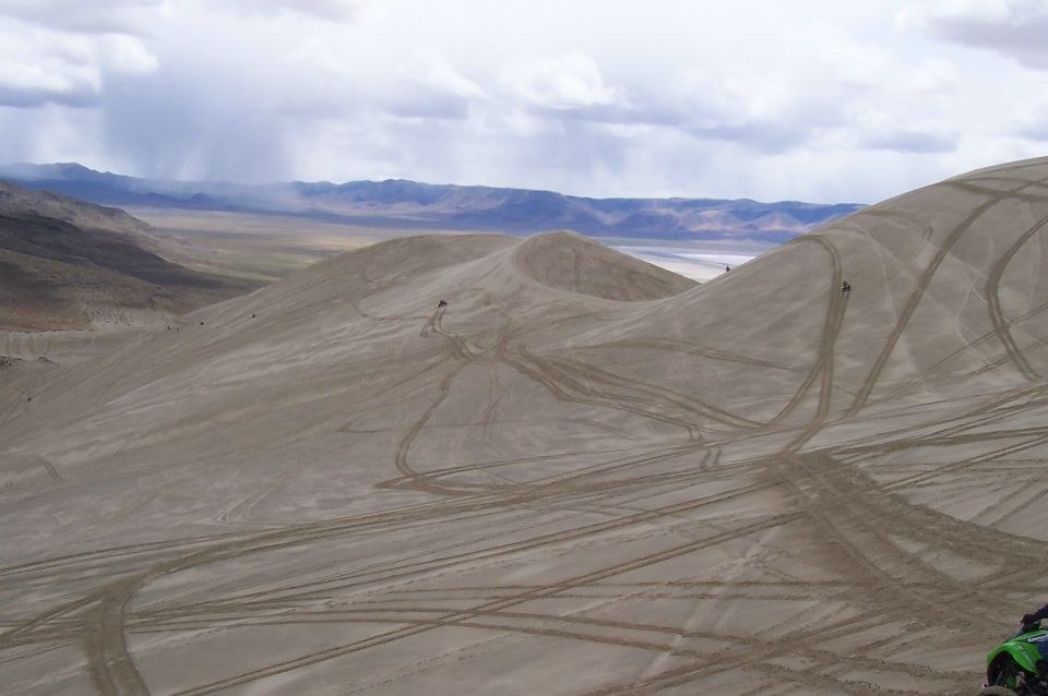 Sand dunes with tire tracks under a cloudy sky, distant mountains visible