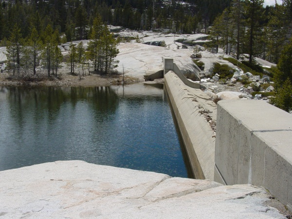 Concrete dam holding back a blue lake, surrounded by granite rocks and pine trees