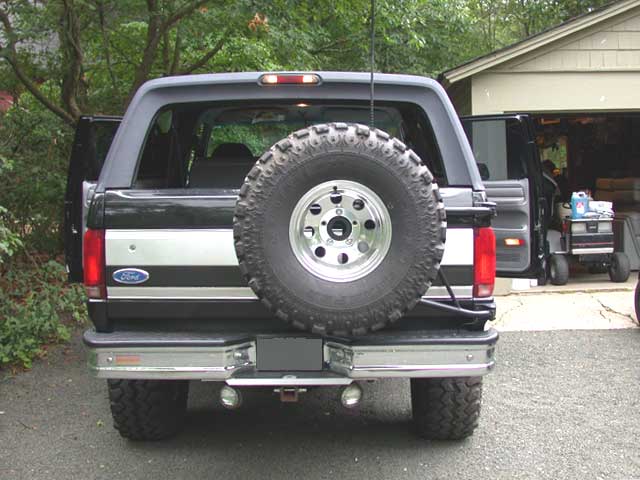 Rear of a lifted black-and-silver Ford Bronco with tailgate spare tire and CB antenna in a driveway