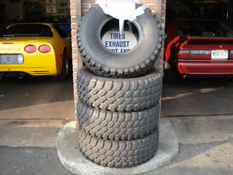 1994 Ford Bronco - A stack of off-road tires sits in front of a garage, partially obscuring a yellow Corvette and a red Mustang.