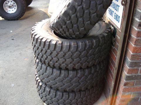 1994 Ford Bronco - A stack of four large off-road tires is shown next to a brick wall, with a sign indicating 'EXHAUST FRONT ENDS'.