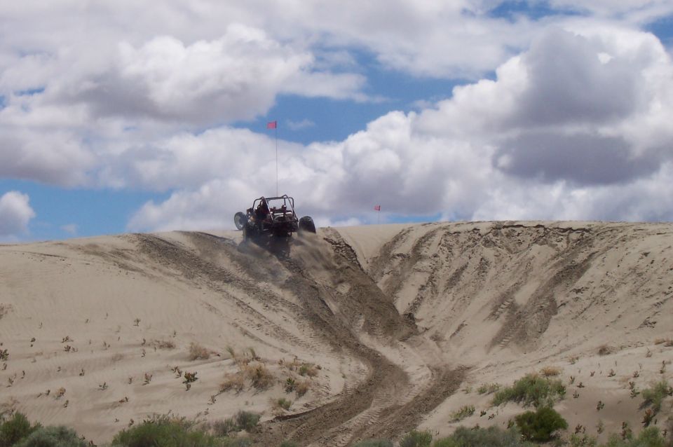 Dune buggy cresting a sand dune with a red flag on top. Dusty trail behind it