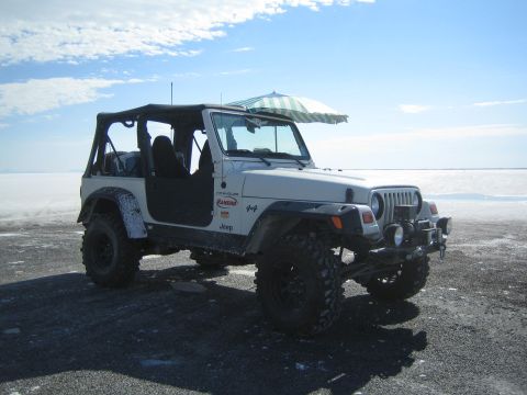 1990 Ford Bronco - white lifted Jeep Wrangler with its doors removed and a beach umbrella sits on a white salt flat under blue sky.