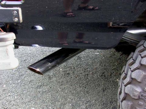 1996 Ford Bronco - Low-angle close-up of a polished chrome exhaust tip with an angle-cut end exiting behind the rear wheel of a black truck, shot over gravel.…