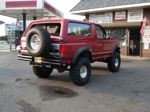 1994 Ford Bronco - Rear three-quarter view of a red lifted Ford Bronco parked outside Hoffman Transmission's Plus auto repair shop, with an uncovered mud-terr…