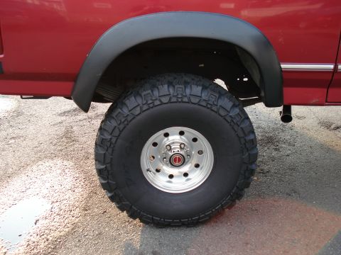 1994 Ford Bronco - Rear wheel close-up on a lifted red Ford truck, with a Dirt Grip mud-terrain tire mounted on a polished slotted alloy wheel wearing a small…