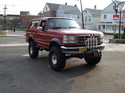 1994 Ford Bronco - Red lifted full-size Ford Bronco photographed from the passenger-side 3/4 front in a paved lot with houses and a Marlboro cigarette price s…