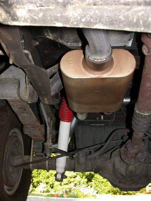 1996 Ford Bronco - Under-vehicle view of a newly installed copper-toned oval muffler with a fresh weld at its inlet pipe, mounted beside a white shock absorbe…