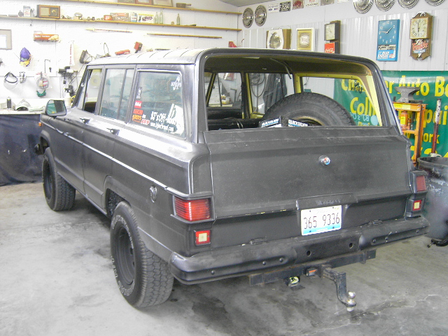 Flat-black Jeep Wagoneer-style SUV in a shop, rear three-quarter view on black steel wheels