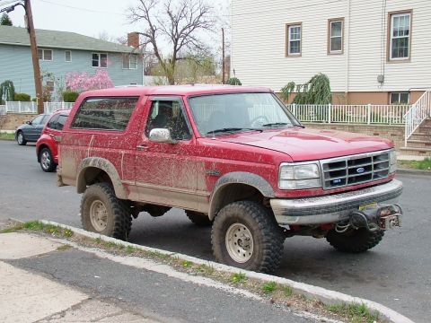 1994 Ford Bronco - Red lifted Ford Bronco splattered with dried mud from the wheels up over the doors and windows, parked along a curb on a residential street…