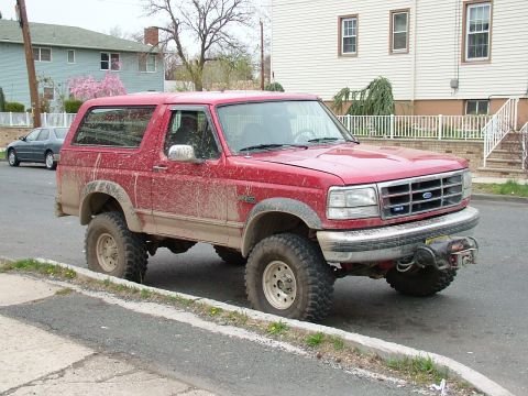 1994 Ford Bronco - Red lifted Ford Bronco caked in dried mud along its flanks and wheels, parked at the curb on a residential street and shot from the front t…