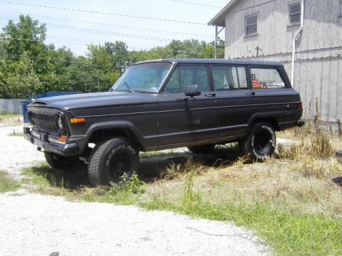 1977 Jeep Wagoneer - A dark gray 1977 Jeep Wagoneer is parked on a gravel lot next to a building.