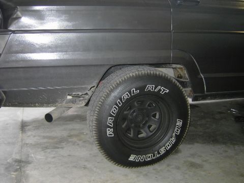 1977 Jeep Wagoneer - The rear passenger side of a Jeep Wagoneer shows significant rust damage near the wheel well and exhaust pipe.