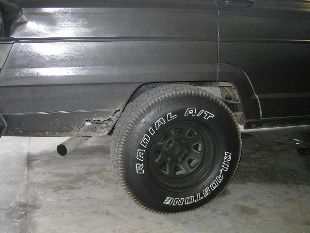 Close-up of a dark-colored Jeep Wagoneer's rear wheel well, showing rust on the lower body panel and a Roadstone Radial A/T tire