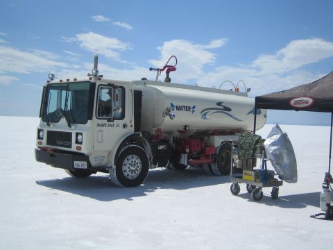 1990 Ford Bronco - white water tanker truck labeled 'WATER' is parked on a white salt flat next to a black pop-up canopy.