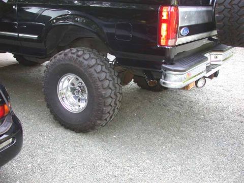 1996 Ford Bronco - Rear quarter close-up of a lifted black Ford Bronco on a gravel driveway, showing an oversized mud-terrain tire on a polished deep-dish all…