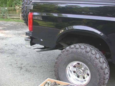 1996 Ford Bronco - Rear quarter of a black lifted Ford truck/Bronco on a gravel driveway, showing the tall Super Swamper mud tire on a polished slotted alloy…