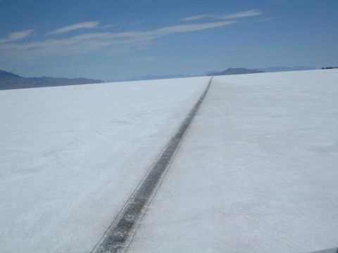 1990 Ford Bronco - lone vehicle track stretches across a bright white salt flat under a clear blue sky with distant mountains.