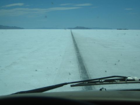1990 Ford Bronco - Driver's-eye view through a windshield of a dark track running across white salt flats toward distant peaks.