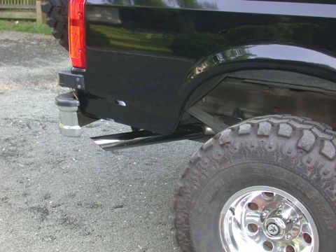 1996 Ford Bronco - Low rear-corner detail of a black Ford SUV showing a chrome side-exit exhaust tip angled out behind the rear wheel, the rear tail light, an…