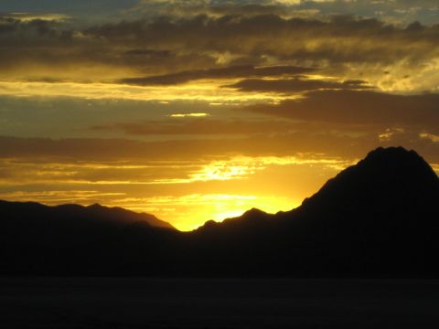 1990 Ford Bronco - vivid golden sunset glows behind the dark jagged silhouette of mountains under cloudy skies.