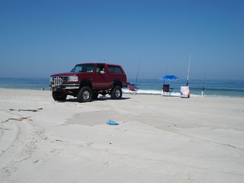 1994 Ford Bronco - Red lifted full-size Ford Bronco with a hardtop parked on wide open beach sand under a clear blue sky, seen from the driver's-side 3/4 fron…