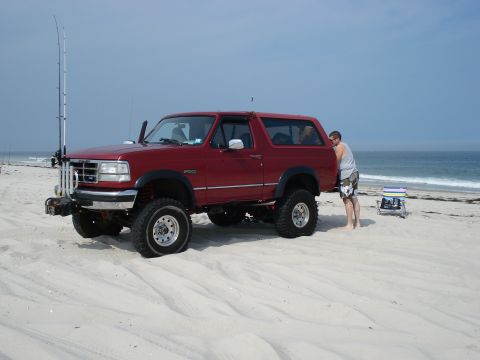 1994 Ford Bronco - Red full-size Ford Bronco with removable hardtop parked on the sand at the ocean's edge, shot from the driver's-side 3/4 front. It runs a s…
