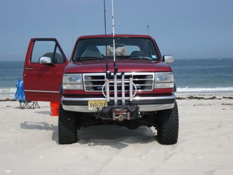 1994 Ford Bronco - Head-on front view of a red lifted Ford Bronco parked facing the ocean on a sand beach, driver's door open and a surf-fishing rod propped a…