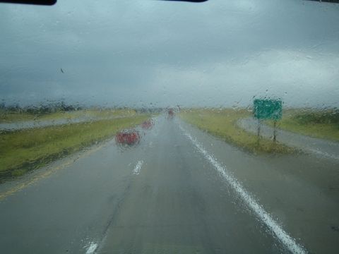 1990 Ford Bronco - View through a rain-streaked windshield of a wet two-lane highway with brake lights and a green road sign ahead.