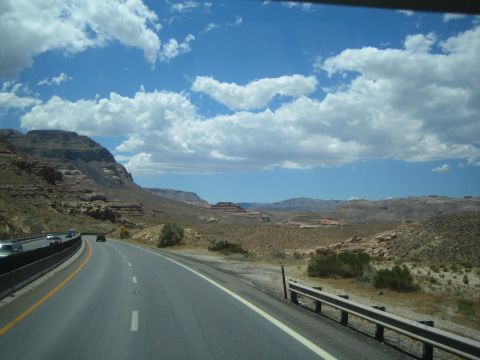 1990 Ford Bronco - highway curves downhill into a broad desert valley dotted with mesas, with traffic ahead under puffy clouds.