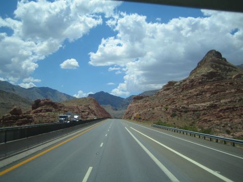 1990 Ford Bronco - highway curves through red and tan rock formations and mountains under a bright blue cloud-dotted sky.