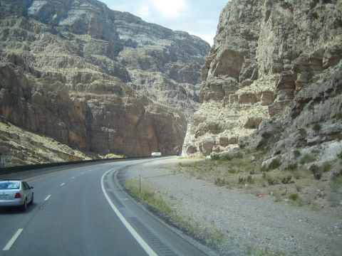 1990 Ford Bronco - curving highway cuts through a steep rocky canyon, a silver sedan in the near lane and an RV ahead.