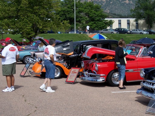 Row of hood-up custom cars at an outdoor show, red 1950s Chevy in front