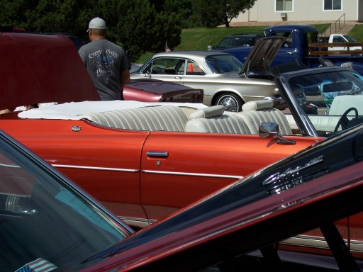 Red-orange convertible with white pleated seats among classics at an outdoor car show