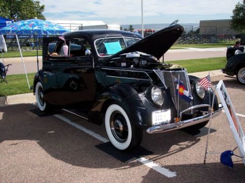 1972 Ford Ranchero - Black 1937 Ford Sedan parked at an outdoor event, hood is open, displaying flags.