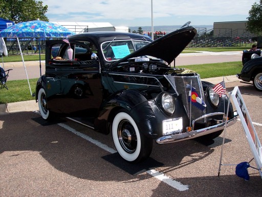 Black 1930s Ford coupe hot rod with hood open and whitewall tires at an outdoor car show